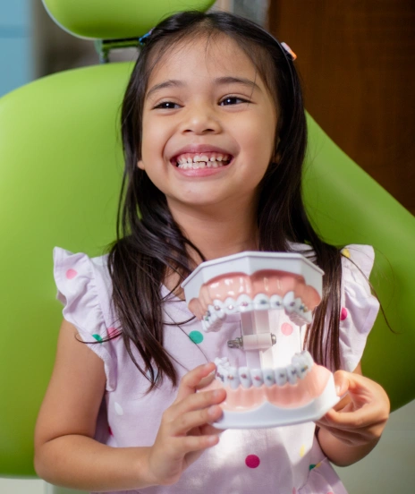 Young girl smiling in dental chair holding orthodontic teeth model demonstrating braces treatment.