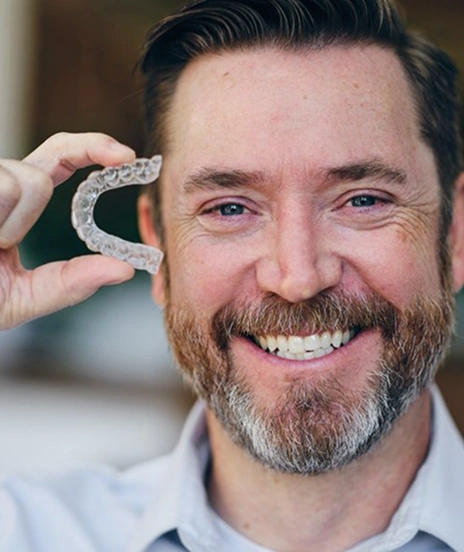 Smiling man with beard holds clear orthodontic aligner near face in professional setting.