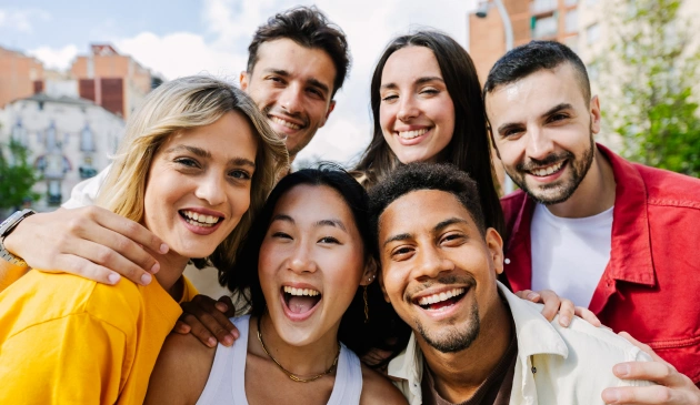Six smiling young people hugging outdoors in front of residential buildings, displaying healthy teeth and happy expressions.