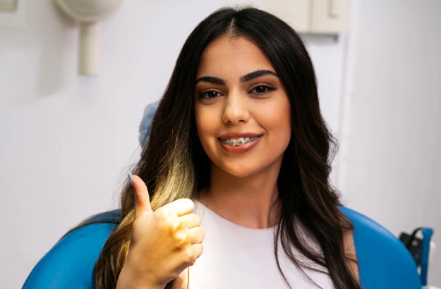 Woman in blue cardigan giving thumbs up in dental office setting, smiling at camera.