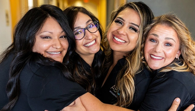 Four smiling women in black clothing posing together indoors for a professional group photo.