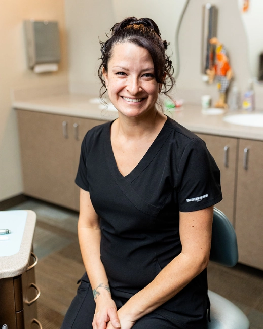 Smiling dental professional in black scrubs seated in modern orthodontic office treatment room.