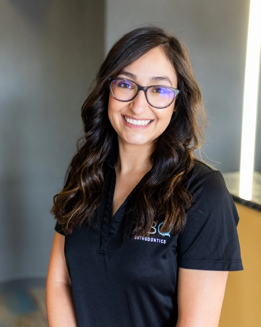 Young woman wearing glasses and black ABQ Orthodontics polo shirt, smiling at camera in professional office setting.