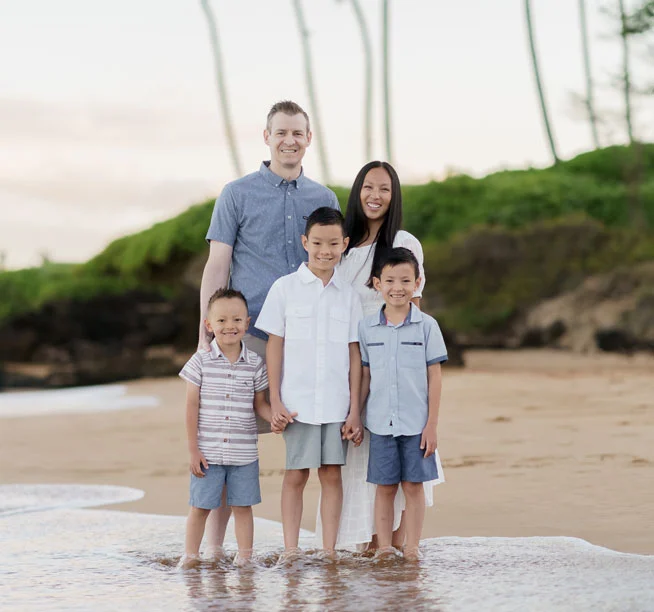 Family of five standing together in shallow ocean water at beach with palm trees in background.